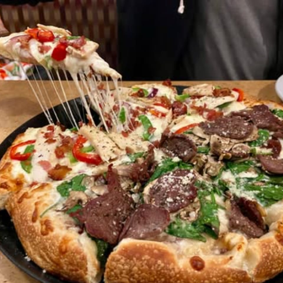 Cheesy meat and vegetable pizza being lifted from a black serving pan, with stretchy melted cheese and a golden crust.