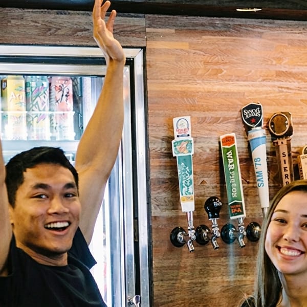Two smiling people, a man with arms raised and a woman, stand in front of beer taps.