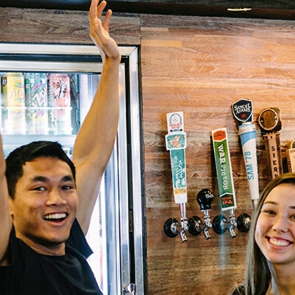 Two smiling people, a man with arms raised and a woman, stand in front of beer taps.