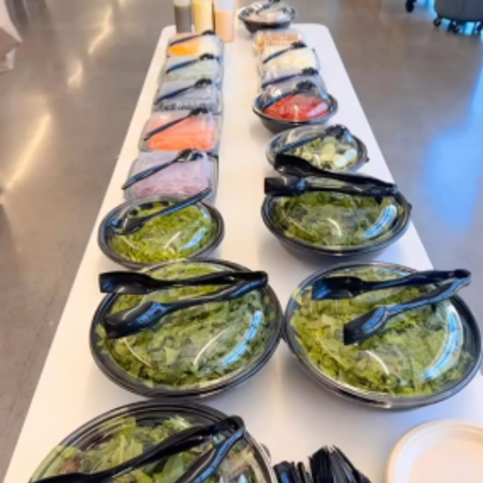 Buffet table displays bowls of green salads with black serving tongs in a bright dining area.