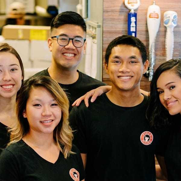 Five smiling young restaurant staff members in black t-shirts with a logo, standing together.