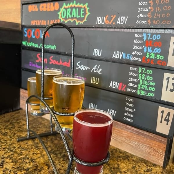 Four glasses of beer, three golden and one red, in a flight holder, with a menu board behind.