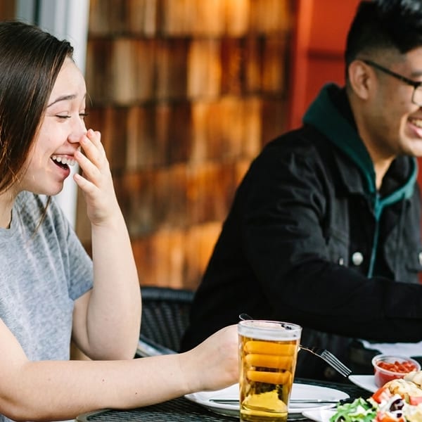 Two people laughing and eating outdoors at a restaurant, with food and drinks on the table.
