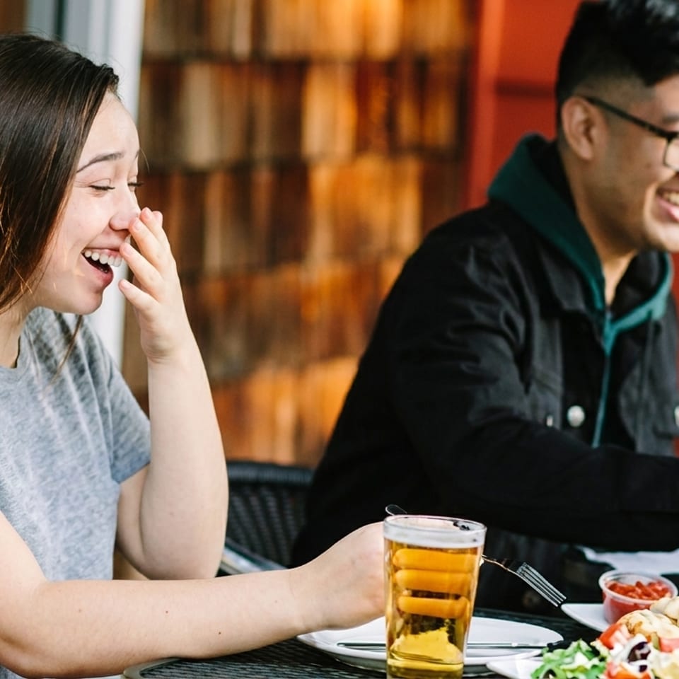 Two people laughing and eating outdoors at a restaurant, with food and drinks on the table.