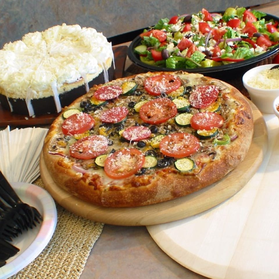 Vegetable pizza topped with tomatoes, zucchini, mushrooms, olives, and cheese beside a fresh salad and cheesy bread.