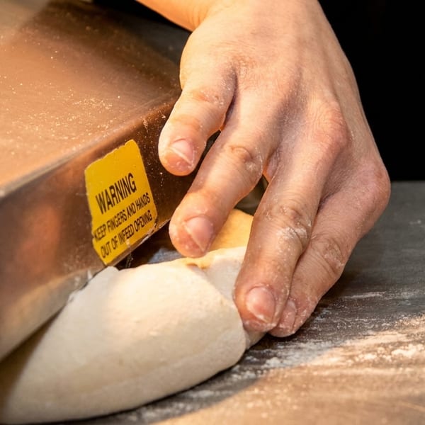 A chef's hands guide dough into a machine with a yellow "WARNING" label.