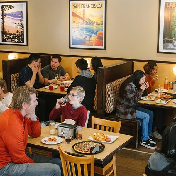 Diverse groups of people enjoy pizza and drinks at tables and booths in a casual restaurant.