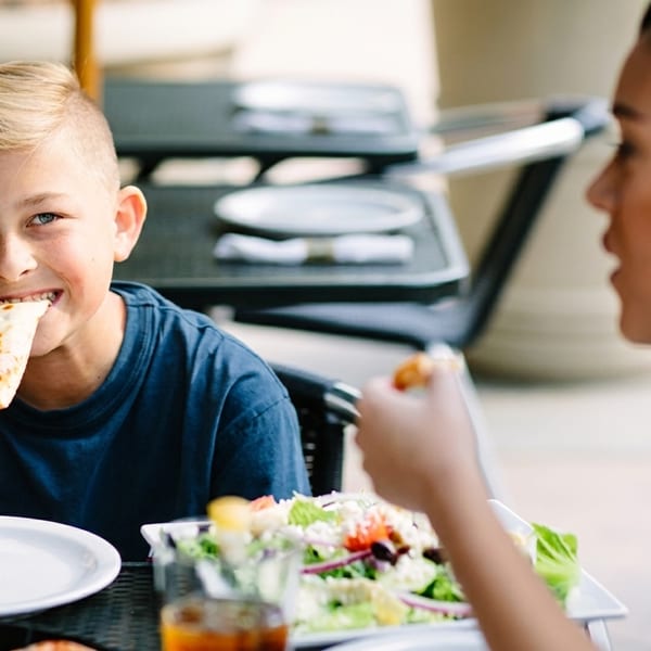 A young boy smiles while eating a slice of pizza at an outdoor restaurant table with an adult.