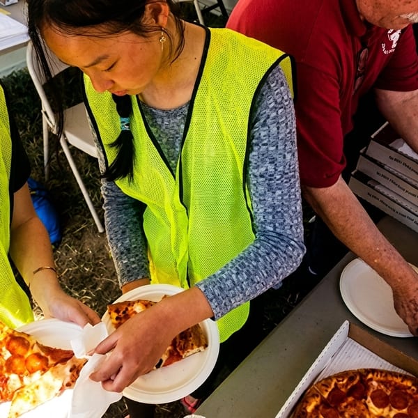 People in safety vests serving slices of pepperoni pizza from boxes outdoors.