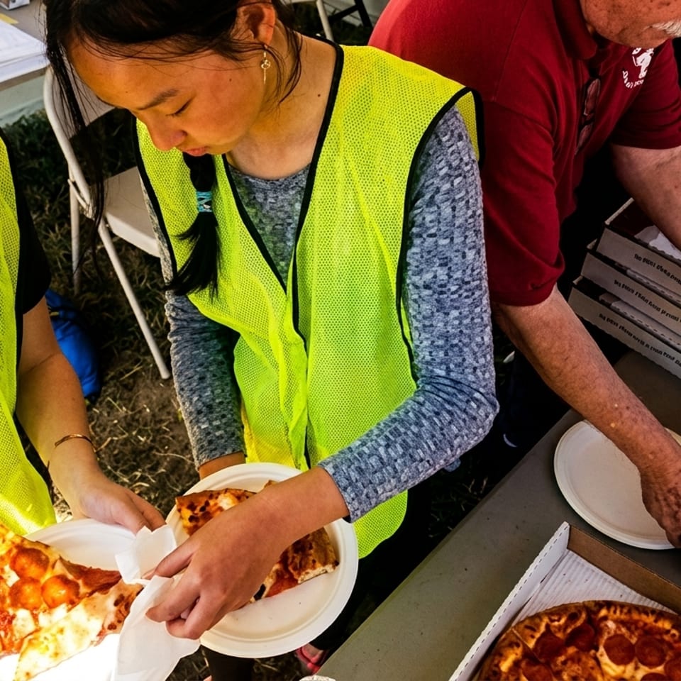 People in safety vests serving slices of pepperoni pizza from boxes outdoors.
