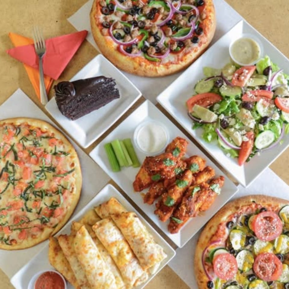 Assorted pizzas, salad, buffalo wings, breadsticks, chocolate cake, and dipping sauces arranged on a dining table.