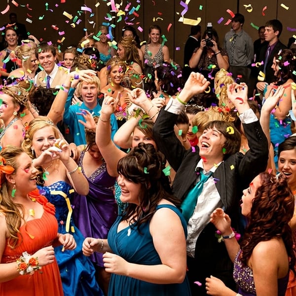 A large group of young people in formal wear celebrate with colorful confetti falling around them.